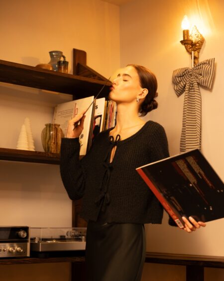 A woman in a cozy tie-front cardigan with minimalist christmas decorations behind her, holding a christmas vinyl and kissing it