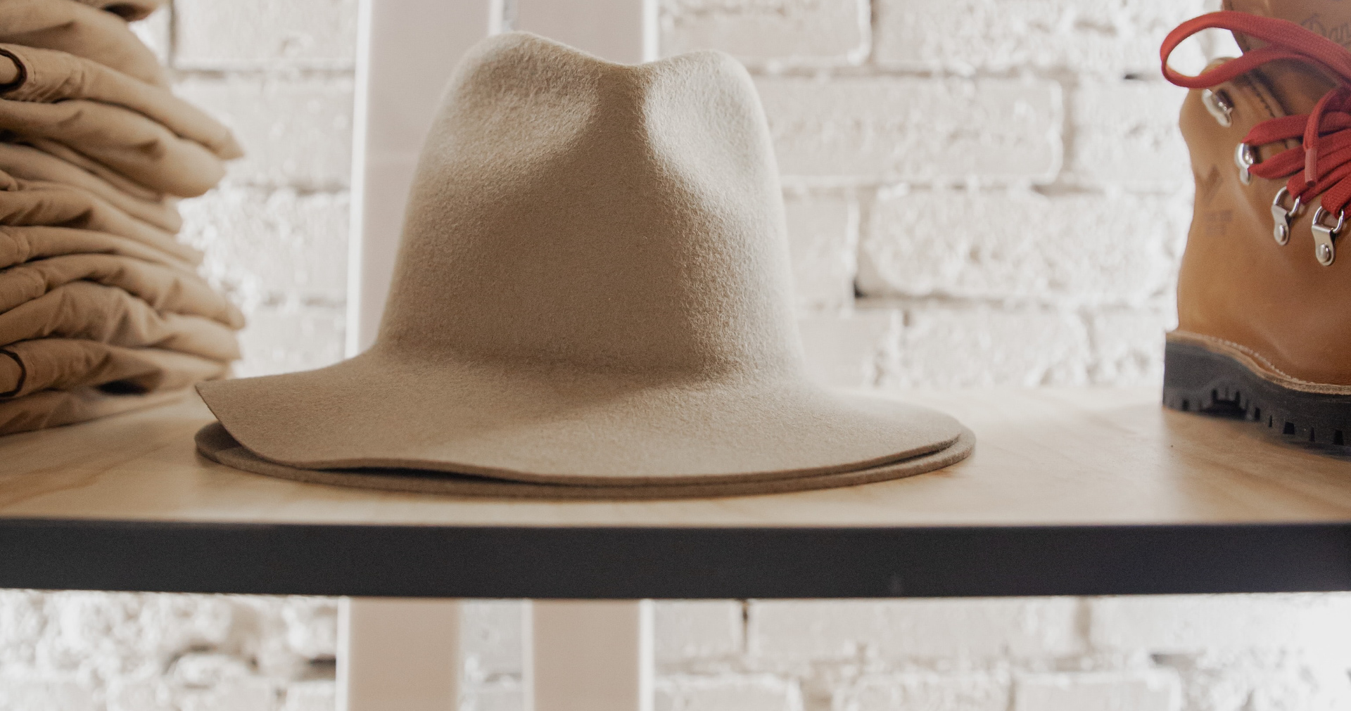 Style Trend Clothiers About Us banner featuring a wooden shelf with beige hats, bulky boots and folded tan pants and a white brick wall in the background