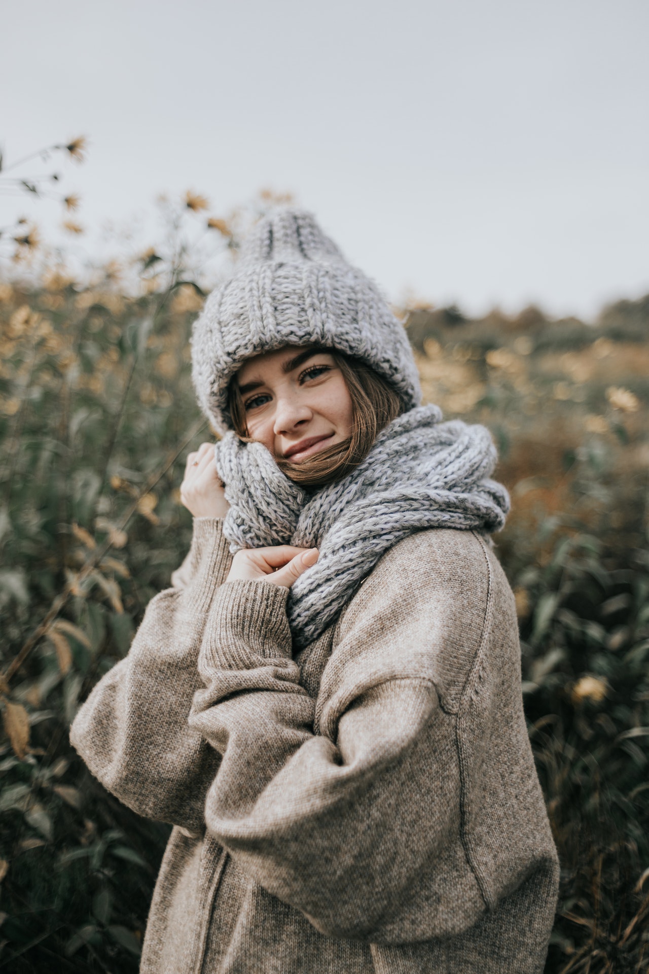 closeup of a woman in a field of flowers in the fall wearing a cozy oversized winter sweater, grey knitted scarf and toque